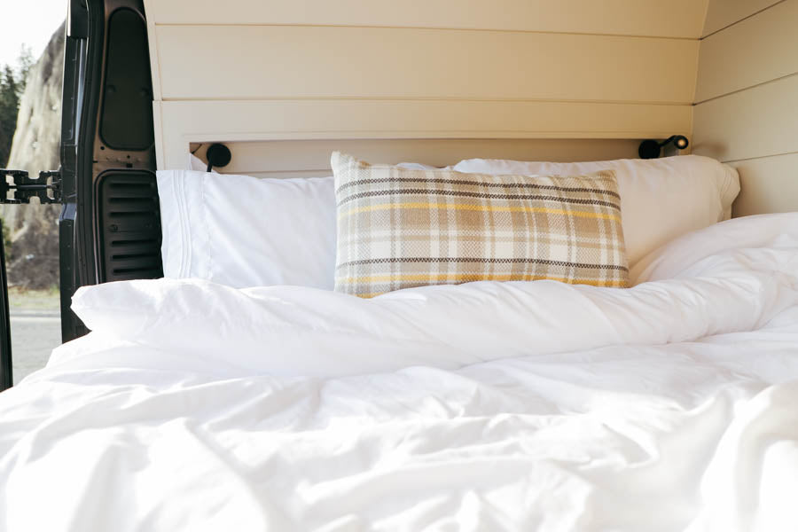 Relax in the inviting bed of our RV rental. Features soft white bedding, a plaid pillow, and outdoor views.