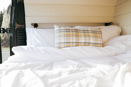 Relax in the inviting bed of our RV rental. Features soft white bedding, a plaid pillow, and outdoor views.