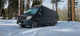 Snow-chained rv rental Sprinter van in a serene winter forest, ready for adventure.