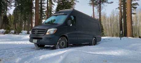 Snow-chained rv rental Sprinter van in a serene winter forest, ready for adventure.
