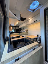 Modern RV rental interior featuring a sleek kitchen with black sink, induction cooktop, light wood counters, and a cozy bed.