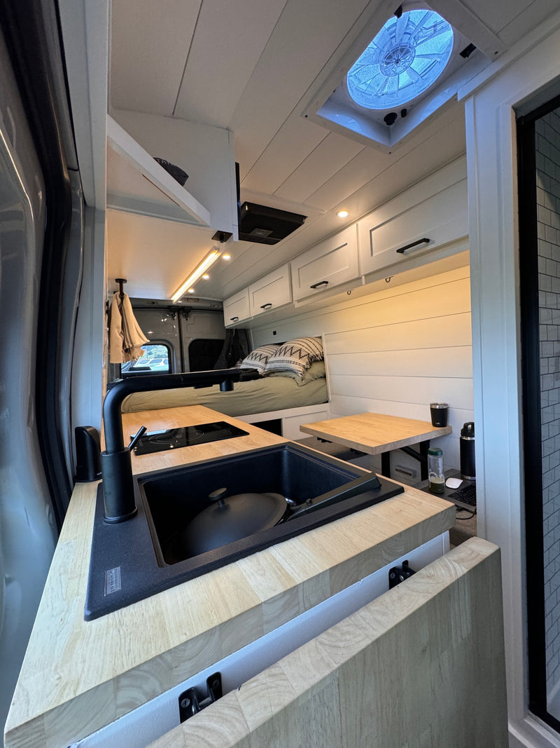 Modern RV rental interior featuring a sleek kitchen with black sink, induction cooktop, light wood counters, and a cozy bed. — photo 5