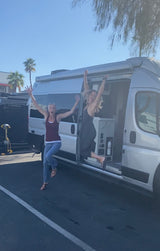 Two excited women joyfully jump next to a silver RV rental van, one inside, one out, celebrating travel.