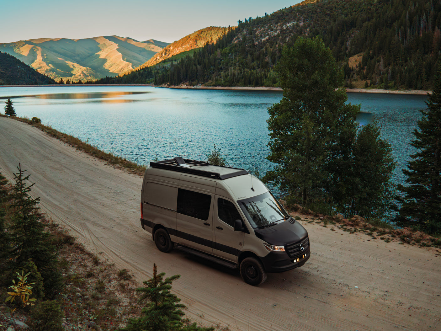 Adventure-ready RV rental camper van parked on a dirt road beside a scenic mountain lake with forests. Perfect for nature exploration.