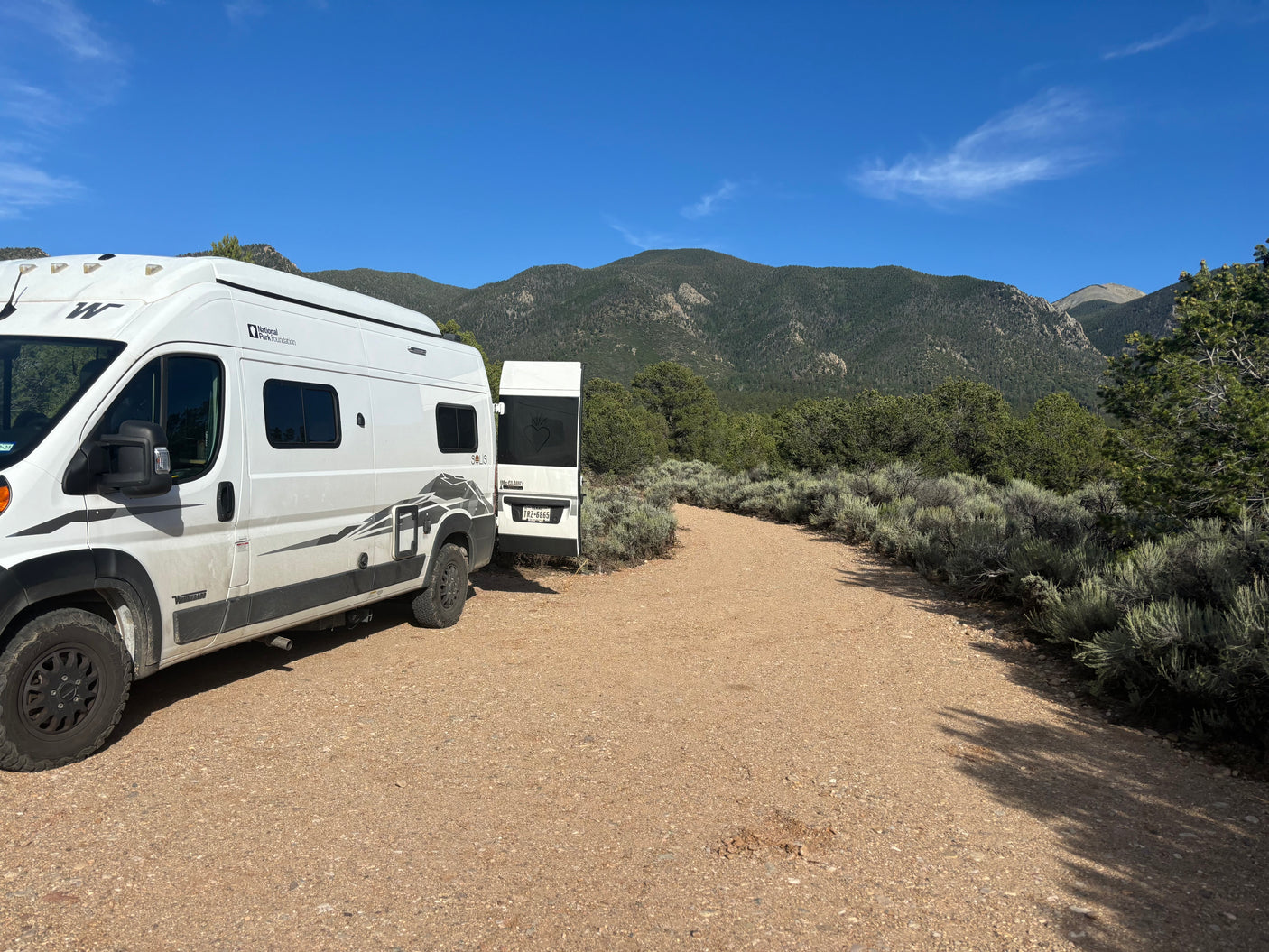 A white Winnebago Solis rv rental parked on a gravel road, surrounded by mountains and sagebrush under a blue sky.