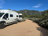 A white Winnebago Solis rv rental parked on a gravel road, surrounded by mountains and sagebrush under a blue sky.