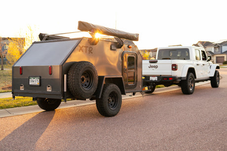 White Jeep Gladiator towing a dark grey XTR off-road teardrop camper, illuminated by warm sunset light for your next RV rental.