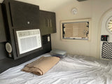 Inside an rv rental: a compact bedroom with a window, striped bed, AC unit, and wood cabinets. Cozy and functional.