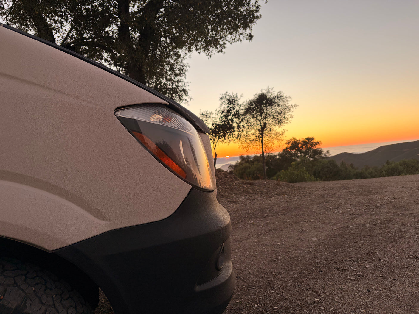 Golden sunset reflects on an RV rental van's headlight on a mountain dirt road, overlooking hazy hills. Perfect for adventure.