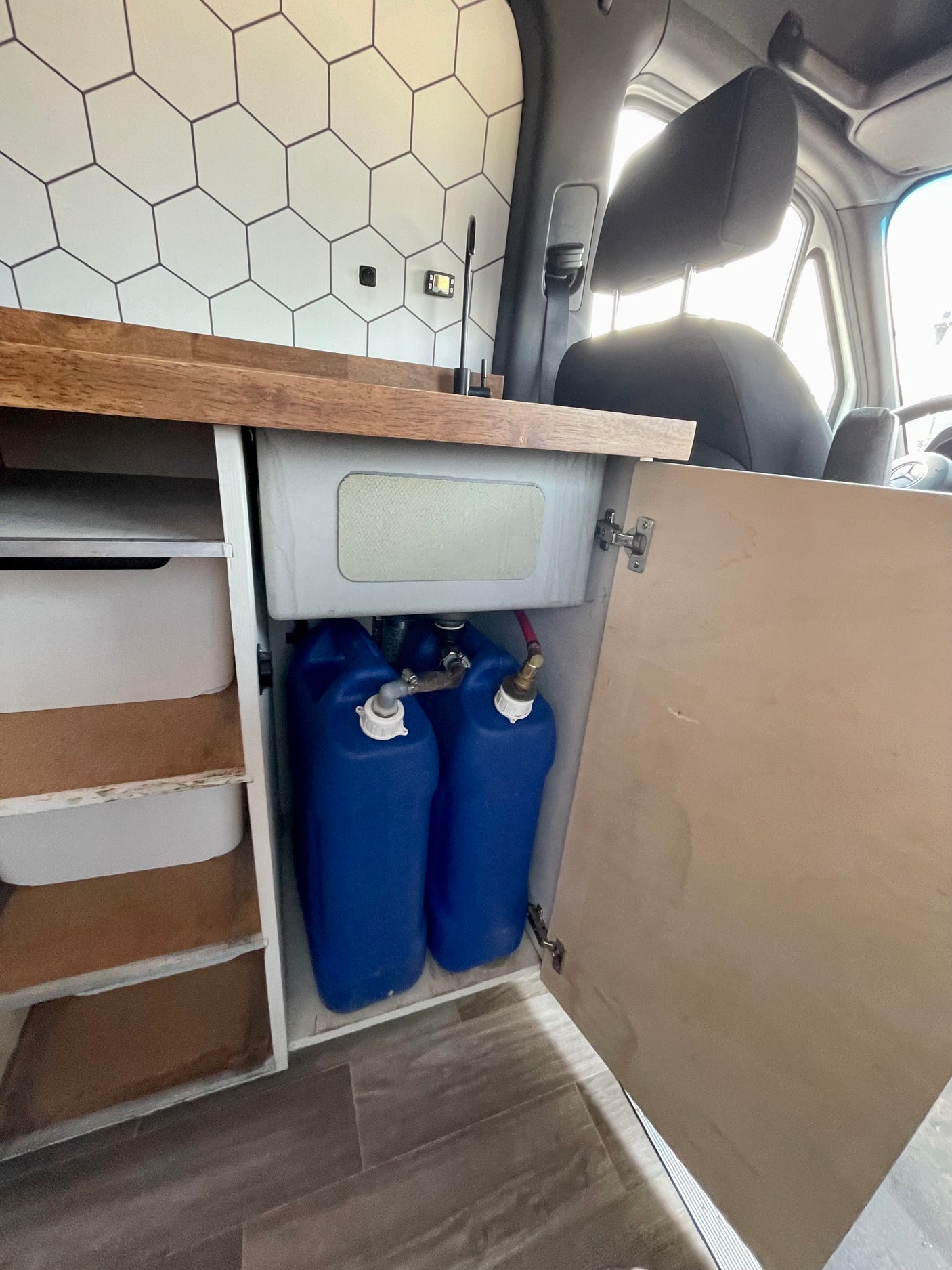 This RV rental interior shows a compact camper van kitchen with a sink, two blue water tanks, and organized storage.