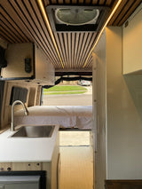 RV rental interior featuring a stylish kitchenette, cozy bed, and custom wood-slatted ceiling with accent lights.