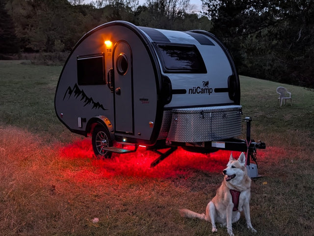 This nüCamp teardrop rv rental with red underglow and a husky mix dog is set for a cozy night of camping adventure.|RV rental: A nüCamp teardrop trailer glows with red underglow lights as a dog sits in a grassy field at dusk.|Inside a clean RV rental