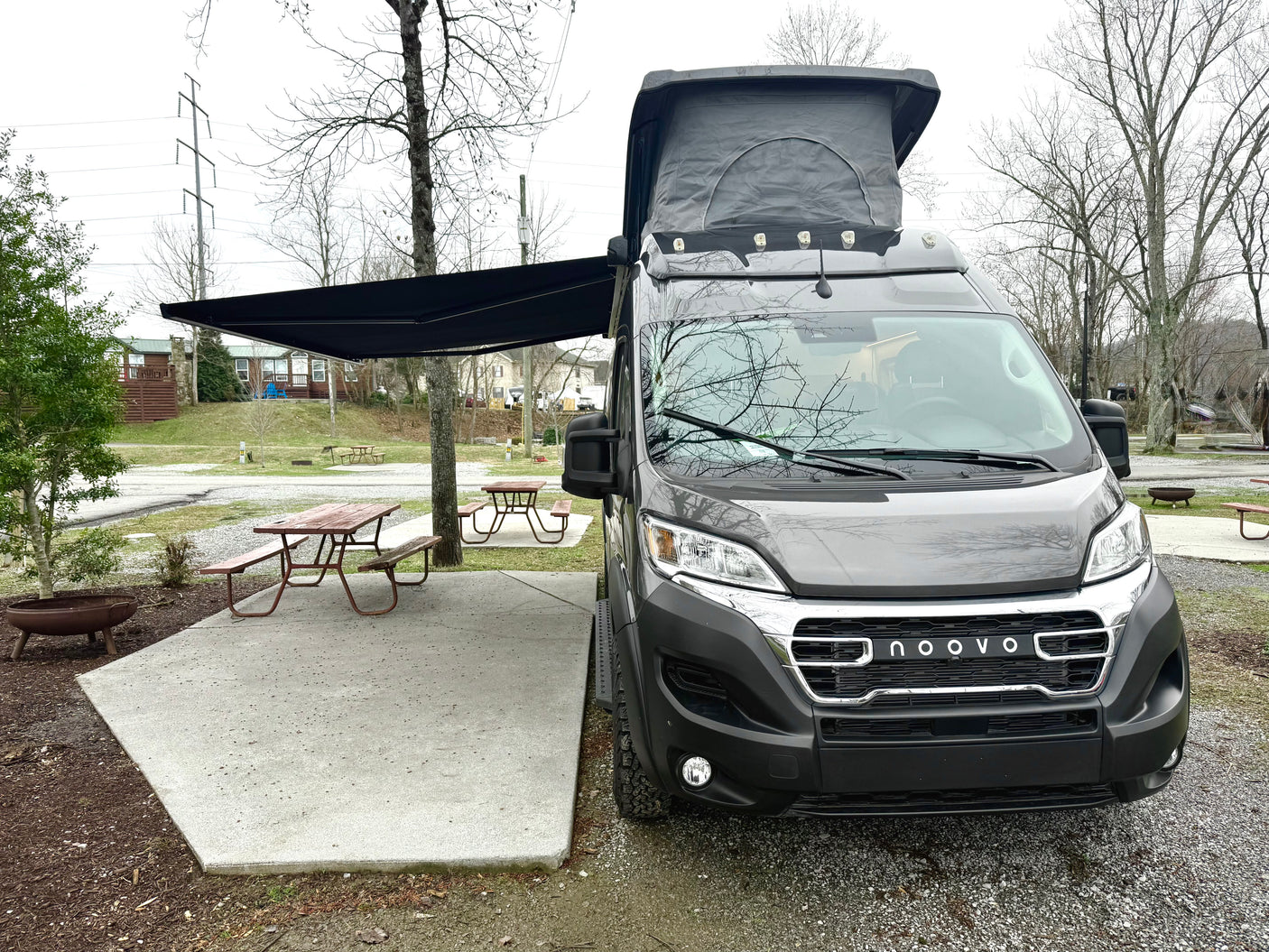 A modern gray Noovo camper van RV rental with pop-top and awning, at a campsite with picnic table.