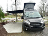 A modern gray Noovo camper van RV rental with pop-top and awning, at a campsite with picnic table.