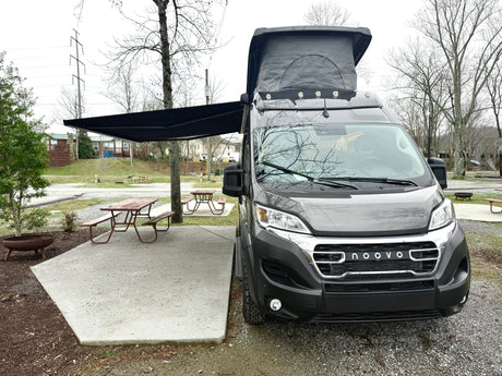 A modern gray Noovo camper van RV rental with pop-top and awning, at a campsite with picnic table.