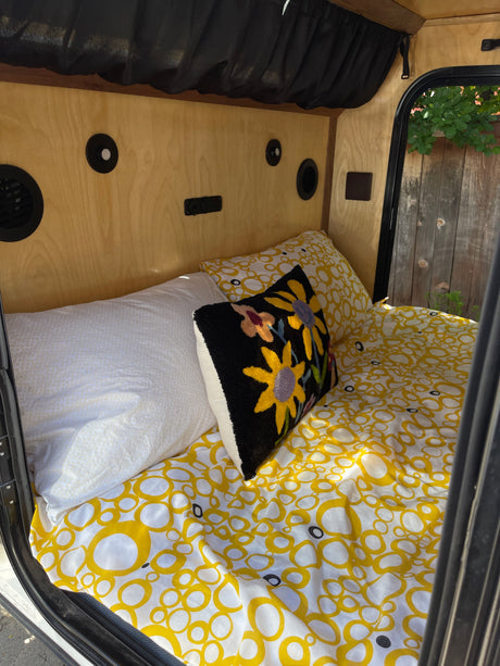 Compact rv rental sleeping area featuring yellow patterned bedding, a colorful floral pillow, and warm wood paneling.