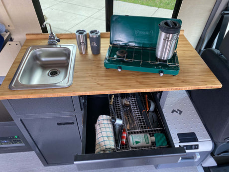 Compact RV rental kitchen with sink, camp stove, cooler, and organized utensils for convenient camp cooking.