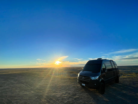 A dark Sprinter van, perfect for an RV rental, is parked in a vast, flat landscape under a glowing sunset.