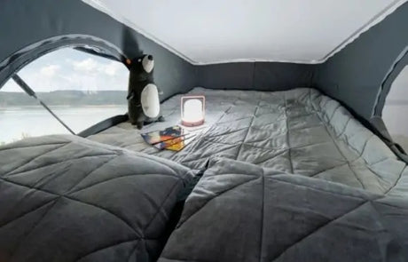 Inside a comfortable RV rental pop-top: a quilted bed, plush toy, glowing lamp, and a stunning lake view.