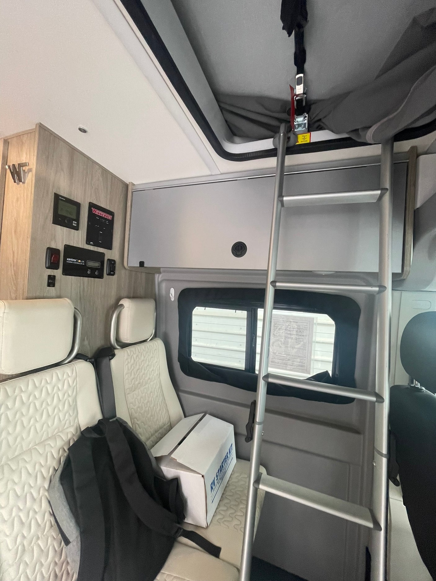Interior of an RV rental, showing a ladder to an upper bunk, control panels on the wall, and an RV starter kit box on patterned seats.