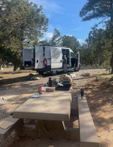 An RV rental camper van parked at a scenic campsite, with a person by the open van and a picnic table set for a meal.