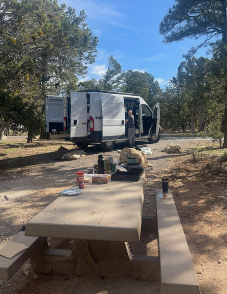 An RV rental camper van parked at a scenic campsite, with a person by the open van and a picnic table set for a meal.