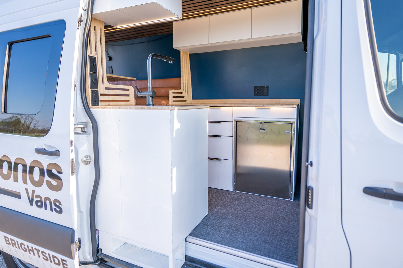 An RV rental: Inside a white camper van, a modern kitchen with a sink, refrigerator, and cabinet storage is visible.