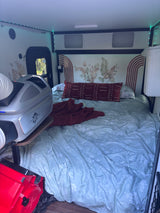 Cozy RV rental interior featuring a bed with botanical decor, LED lighting, and a portable Zero Breeze AC unit for comfort.