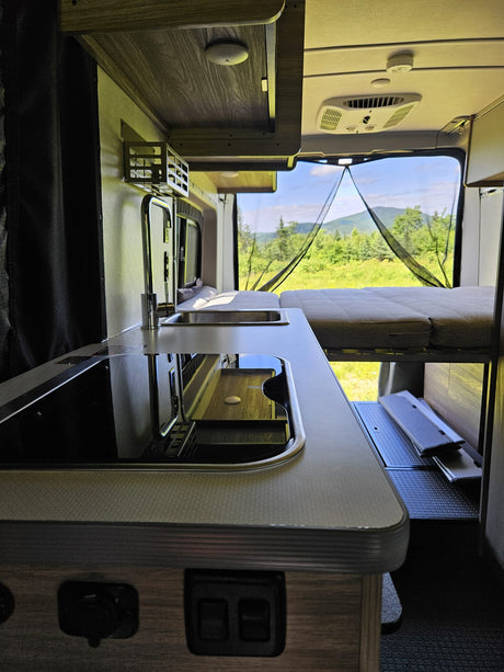 Inside an RV rental: modern kitchenette, elevated bed, and a mosquito-netted rear view of mountains and a lush landscape.