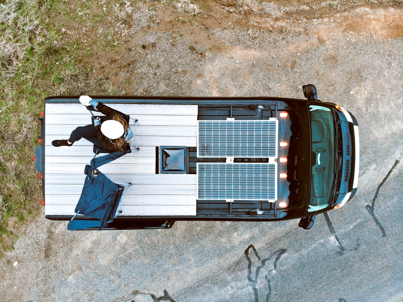 Top-down view of a black camper van with solar panels. A person enjoys the roof deck. Perfect for an RV rental adventure!