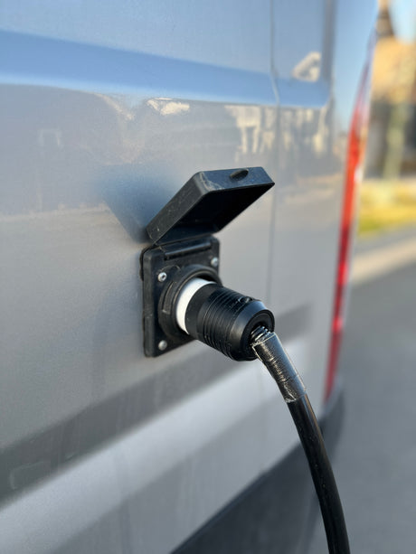 This RV rental is connected to shore power. A black electrical cord is plugged into the gray van's outdoor inlet.
