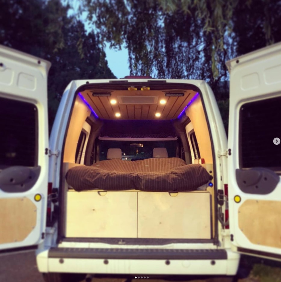  offering fresh air and comfort.|An RV rental's interior reveals a cozy pinstriped bed and a practical wood panel with a power switch & 12V outlets.|RV rental charging ports: USB-C PD