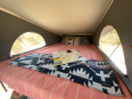 Inside a pop-top RV rental: a comfy bed with map, binoculars, and blankets. Large windows show nature.