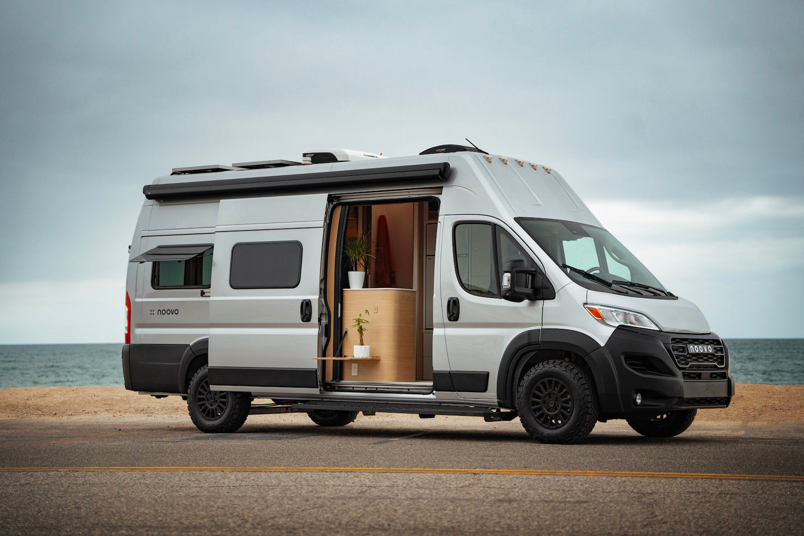 Silver Noovo camper van RV rental parked by the ocean with its door open, revealing a modern, plant-filled interior.