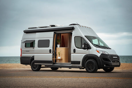 Silver Noovo camper van RV rental parked by the ocean with its door open, revealing a modern, plant-filled interior.