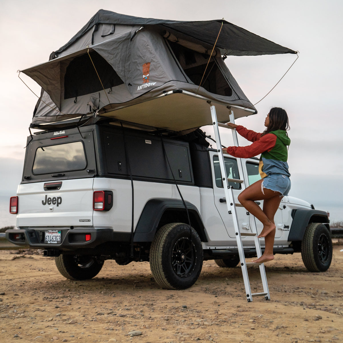 Adventure awaits! Rent this white Jeep Gladiator RV rental with a rooftop tent. Woman climbing ladder for camping.
