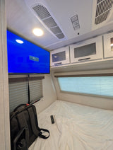 Inside an RV rental bedroom: A mounted TV, AC unit, overhead cabinets, and a bed with a remote and backpack.