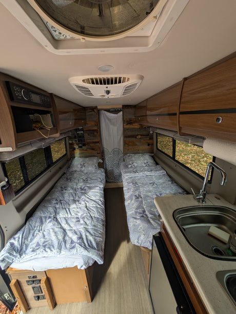 Inside this cozy rv rental: twin beds with patterned comforters, a compact kitchen with sink, wood walls, and overhead storage.