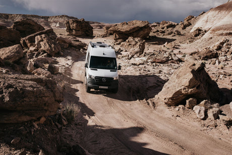 White Mercedes RV rental van drives on a dirt road in a vast, rocky desert with large boulders under a dramatic sky.