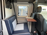 Compact rv rental interior showing blue & white seating, a window, control panel, and a laptop on a wooden table.