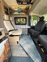 Modern camper van interior for RV rental, featuring swiveling seats, table, and scenic wall art. Cozy and functional.