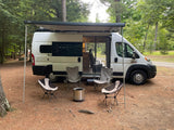 RV rental camper van with awning, chairs, and Solo Stove fire pit set up at a forest campsite. Perfect for outdoor adventures.