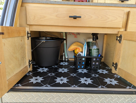RV rental under-counter storage reveals a black bucket, cleaning supplies in baskets, and distinctive star-patterned flooring. Organized for travel.