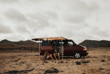 Relaxing rv rental adventure: A couple unwinds in front of their maroon camper van with lights, parked on sandy terrain near mountains.