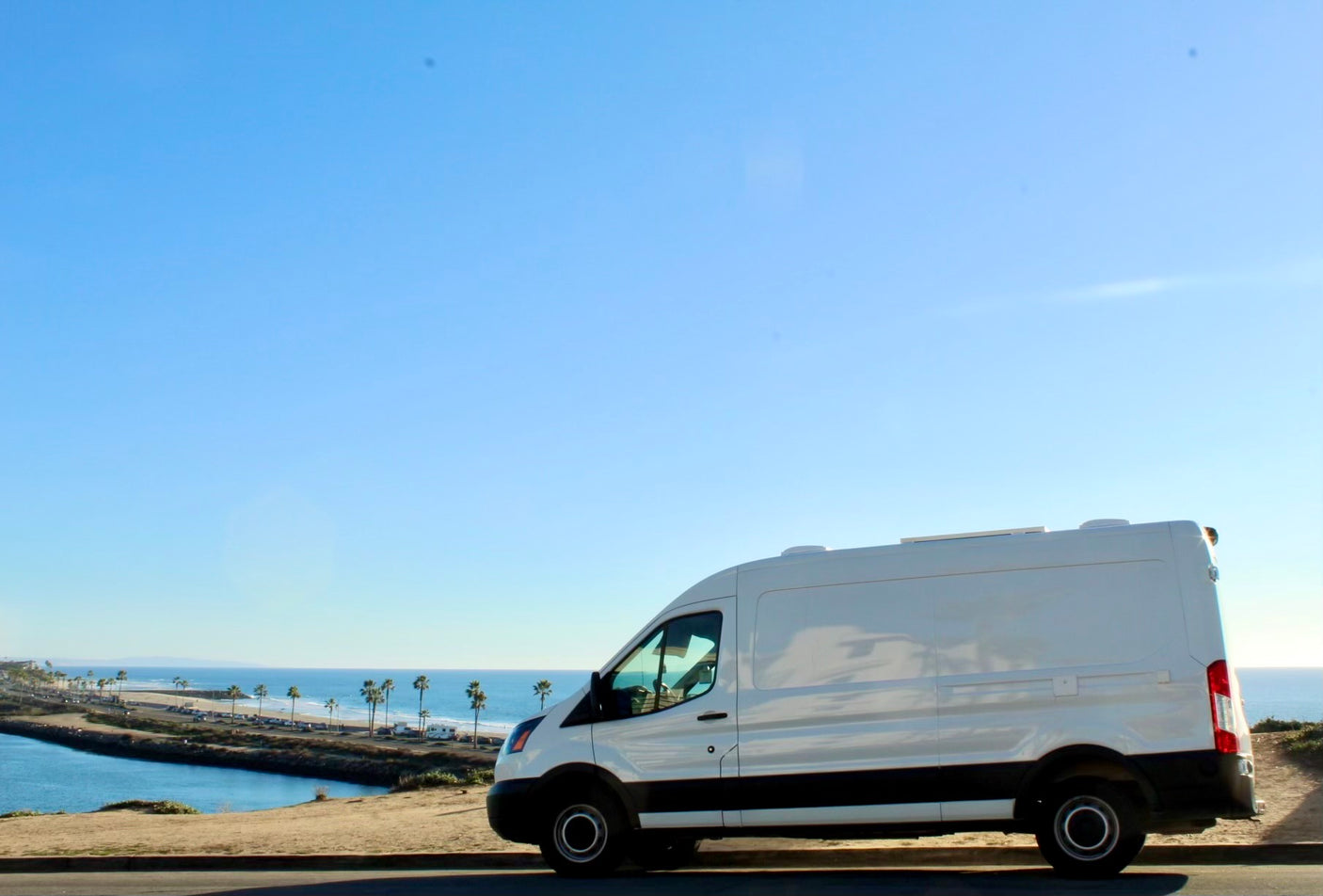 White van (RV rental) parked on a coastal road, overlooking ocean, palm trees, and a channel under a clear sky.
