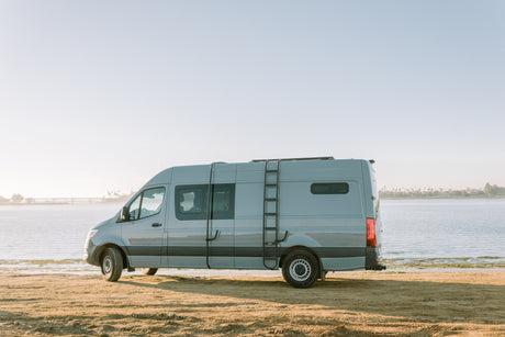 Experience freedom with this RV rental. A grey camper van overlooks a calm waterfront from a sandy beach.