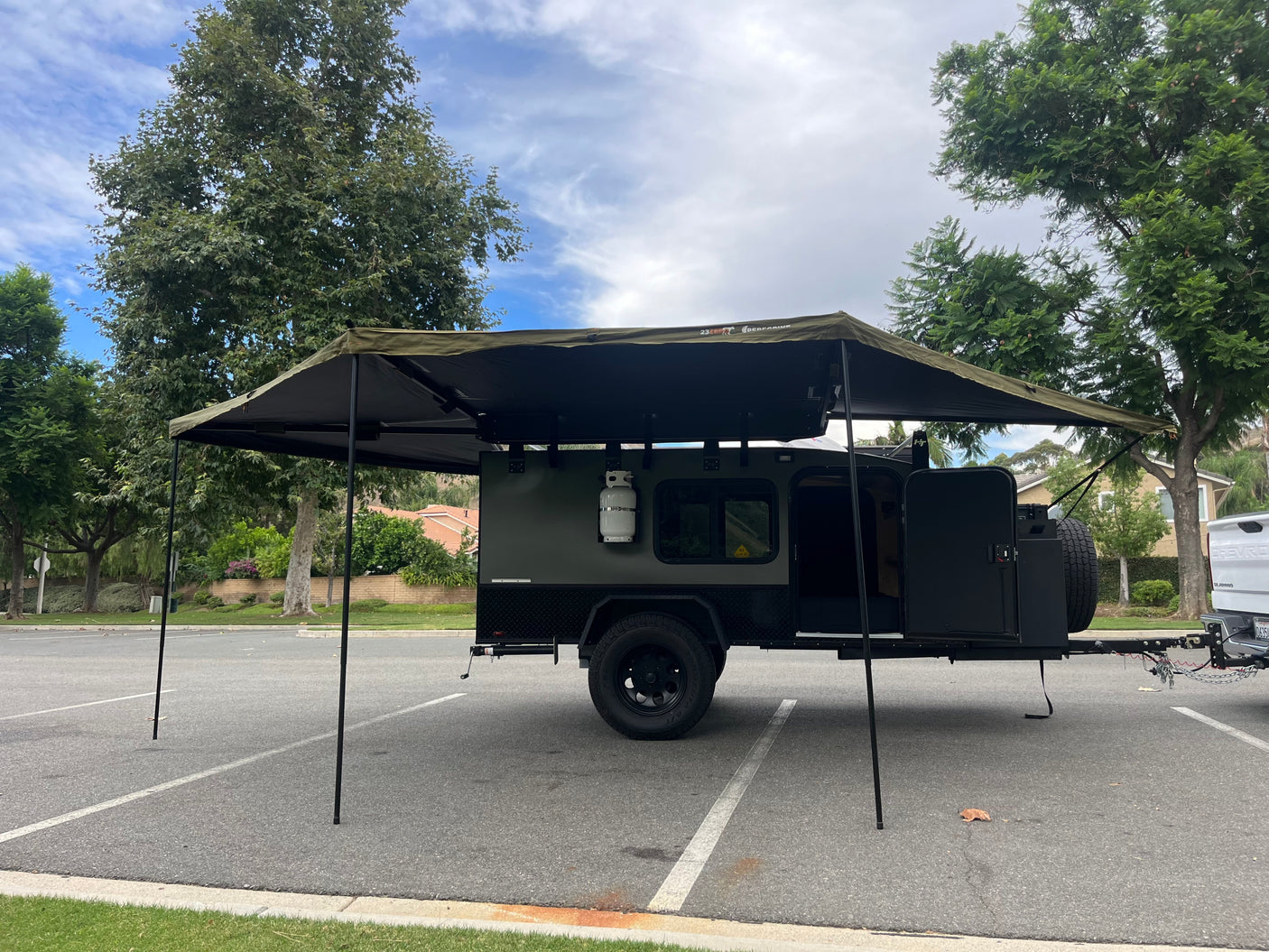 Dark green off-road camper trailer with extended awning and visible propane tank, resembling an RV rental, parked in a lot.