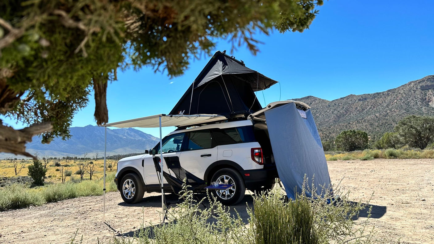 White SUV with rooftop tent & side awning parked in a desert landscape with mountains. A compact RV rental alternative.