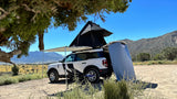 White SUV with rooftop tent & side awning parked in a desert landscape with mountains. A compact RV rental alternative.