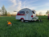 Cozy teardrop RV rental in a green field with an open hatch, side window, and a bright campfire at sunset.